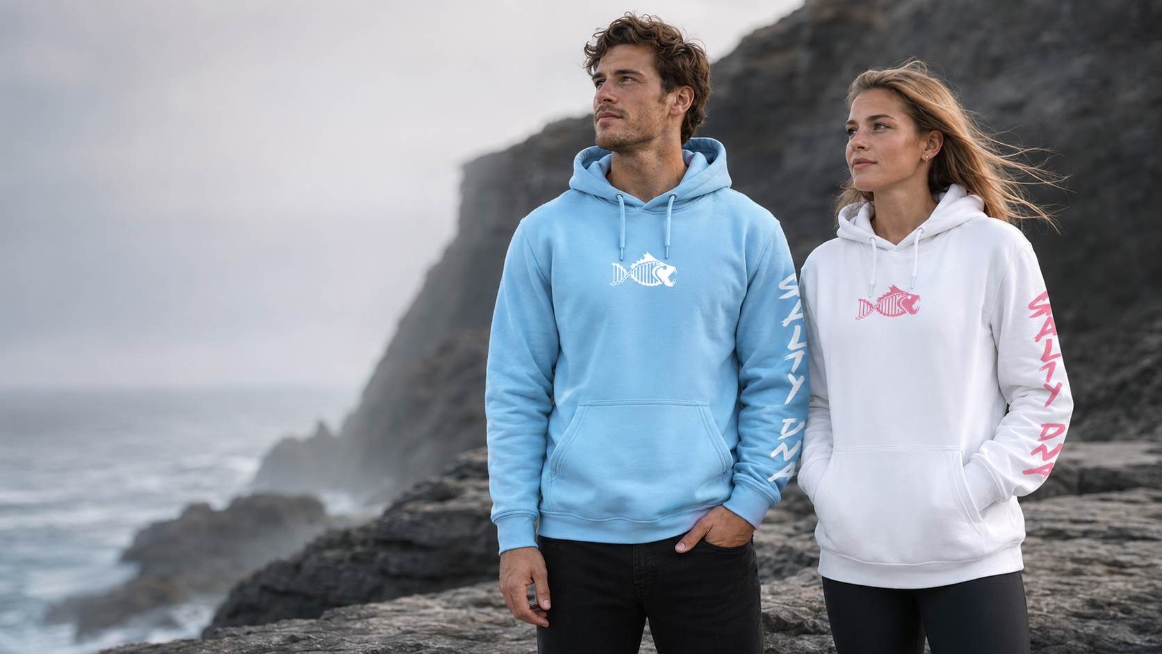 A man and a woman standing on a rocky cliffside by the ocean, wearing matching hoodie sweatshirts. The man wears a light blue hoodie with a white fish logo on the chest and white sleeve graphics, while the woman wears a white hoodie with a pink fish logo on the chest and pink sleeve graphics.