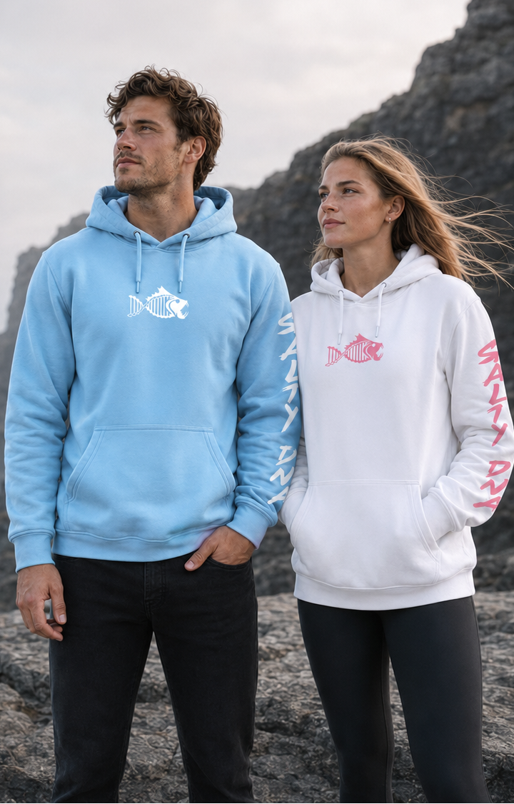A man and a woman standing on a rocky coastal cliffside under an overcast sky, wearing matching hoodies. The man wears a light blue hoodie with a white fish logo on the chest, and the woman wears a white hoodie with a pink fish logo on the chest and pink text down the sleeve.